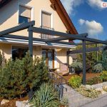 Zdjęcie przedstawia nowoczesną pergolę aluminiową Solid w wersji przyściennej, zamontowaną nad tarasem domu jednorodzinnego. Konstrukcja w kolorze ciemnego antracytu posiada dach tkaninowy, który na tym ujęciu jest całkowicie zwinięty i ukryty pod estetycznym daszkiem ochronnym przy elewacji. Budynek charakteryzuje się jasną, beżową elewacją, brązową dachówką oraz oknami wyposażonymi w zewnętrzne rolety. Taras jest podniesiony, prowadzą do niego kamienne schody, a całe otoczenie wypełnia starannie urządzona zieleń, w tym okazałe krzewy iglaste i ozdobna jukka. Wzdłuż ścieżki na pierwszym planie widoczne są nowoczesne lampy ogrodowe w formie słupków.