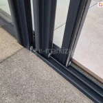 Prowadnica dolna systemu szyb przesuwnych Sotro ramowych w kolorze antracytowym zamocowana do płyty betonowej. Na prowadnicy widoczne trzy skrzydła drzwiowe w kolorze prowadnicy z uszczelkami szczotkowymi. W tle za prowadnicą dodatkowe płytki, którymi wyłożono taras.
