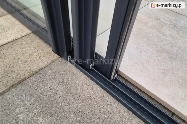 Prowadnica dolna systemu szyb przesuwnych Sotro ramowych w kolorze antracytowym zamocowana do płyty betonowej. Na prowadnicy widoczne trzy skrzydła drzwiowe w kolorze prowadnicy z uszczelkami szczotkowymi. W tle za prowadnicą dodatkowe płytki, którymi wyłożono taras.