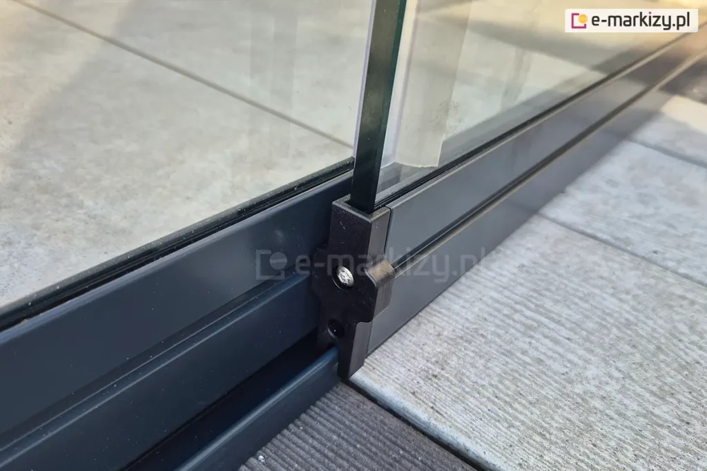 Panel szklanych drzwi przesuwnych bezramowych Miros z zamocowanym na końcu profilem z elementami jezdnymi ukrytymi pod aluminiową osłoną w kolorze antracytowym. Prowadnica dolna zamocowana na tarasie z desek.