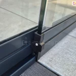 Panel szklanych drzwi przesuwnych bezramowych Miros z zamocowanym na końcu profilem z elementami jezdnymi ukrytymi pod aluminiową osłoną w kolorze antracytowym. Prowadnica dolna zamocowana na tarasie z desek.