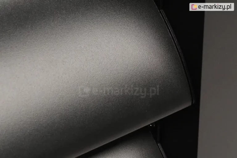 Zbliżenie na strukturę powierzchni aluminiowego profilu lamelowego w kolorze antracytowym, charakteryzującego się matowym, drobnoziarnistym wykończeniem typu struktura. Zdjęcie prezentuje krawędź wygiętego elementu dachu żaluzjowego, na którym widoczne są delikatne refleksy światła podkreślające teksturę lakieru proszkowego. W tle, po prawej stronie, znajduje się pionowy profil nośny, a cała kompozycja ukazuje wysoką jakość wykończenia detali technicznych systemu paneli żaluzjowych Louvre.