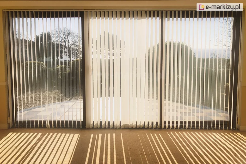 Szerokie przeszklenie tarasowe w salonie, osłonięte przez żaluzje pionowe o białych, lekko transparentnych lamelach. System składa się z trzech sekcji zamontowanych w czarnych ramkach okiennych, które sięgają od sufitu do samej podłogi. Żaluzje pionowe są ustawione pod kątem, co pozwala na przenikanie naturalnego światła do wnętrza, jednocześnie zapewniając prywatność. Przez prześwity między lamelami widoczny jest przydomowy taras wyłożony jasnymi płytkami oraz ogród z krzewami i drzewami w pełnym słońcu. Na beżowej wykładzinie dywanowej wewnątrz pomieszczenia światło słoneczne tworzy wyraźny, rytmiczny wzór długich, równoległych cieni, które podkreślają geometrię osłon okiennych. Konstrukcja jest wykończona dyskretną białą szyną górną, a u dołu lamele połączone są klasycznym białym łańcuszkiem koralikowym, który stabilizuje ich pozycję. Całość aranżacji emanuje spokojem i nowoczesną funkcjonalnością, typową dla nasłonecznionych stref wypoczynkowych.