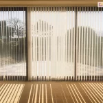 Szerokie przeszklenie tarasowe w salonie, osłonięte przez żaluzje pionowe o białych, lekko transparentnych lamelach. System składa się z trzech sekcji zamontowanych w czarnych ramkach okiennych, które sięgają od sufitu do samej podłogi. Żaluzje pionowe są ustawione pod kątem, co pozwala na przenikanie naturalnego światła do wnętrza, jednocześnie zapewniając prywatność. Przez prześwity między lamelami widoczny jest przydomowy taras wyłożony jasnymi płytkami oraz ogród z krzewami i drzewami w pełnym słońcu. Na beżowej wykładzinie dywanowej wewnątrz pomieszczenia światło słoneczne tworzy wyraźny, rytmiczny wzór długich, równoległych cieni, które podkreślają geometrię osłon okiennych. Konstrukcja jest wykończona dyskretną białą szyną górną, a u dołu lamele połączone są klasycznym białym łańcuszkiem koralikowym, który stabilizuje ich pozycję. Całość aranżacji emanuje spokojem i nowoczesną funkcjonalnością, typową dla nasłonecznionych stref wypoczynkowych.