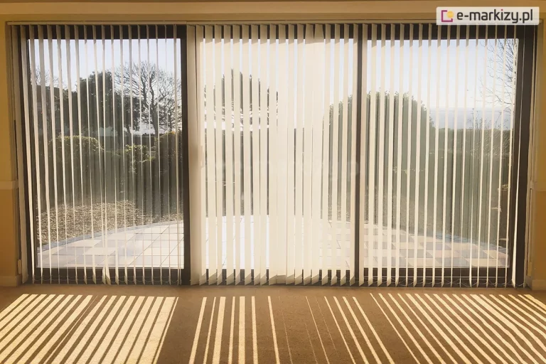 Szerokie przeszklenie tarasowe w salonie, osłonięte przez żaluzje pionowe o białych, lekko transparentnych lamelach. System składa się z trzech sekcji zamontowanych w czarnych ramkach okiennych, które sięgają od sufitu do samej podłogi. Żaluzje pionowe są ustawione pod kątem, co pozwala na przenikanie naturalnego światła do wnętrza, jednocześnie zapewniając prywatność. Przez prześwity między lamelami widoczny jest przydomowy taras wyłożony jasnymi płytkami oraz ogród z krzewami i drzewami w pełnym słońcu. Na beżowej wykładzinie dywanowej wewnątrz pomieszczenia światło słoneczne tworzy wyraźny, rytmiczny wzór długich, równoległych cieni, które podkreślają geometrię osłon okiennych. Konstrukcja jest wykończona dyskretną białą szyną górną, a u dołu lamele połączone są klasycznym białym łańcuszkiem koralikowym, który stabilizuje ich pozycję. Całość aranżacji emanuje spokojem i nowoczesną funkcjonalnością, typową dla nasłonecznionych stref wypoczynkowych.