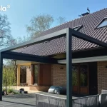 Zdjęcie prezentuje nowoczesną, prostokątną pergolę tarasową Tarasola Flat o konstrukcji aluminiowej w kolorze antracytowym, zamontowaną jako zadaszenie przestronnego tarasu wykonanego z ciemnych desek kompozytowych. Konstrukcja o minimalistycznym designie jest zintegrowana z elewacją parterowego budynku wykonanego z jasnobrązowej cegły klinkierowej i przykrytego spadzistym dachem z ciemnej dachówki. Pod pergolą znajduje się zestaw mebli wypoczynkowych z szarego technorattanu lub sznurka, składający się z narożnika z miękkimi poduszkami. Dach tkaninowy pergoli w kolorze białym jest obecnie całkowicie zwinięty w stronę elewacji, co odsłania widok na niebo i wpuszcza pełne światło na taras. W tle widoczny jest zadbany, zielony ogród z dużymi drzewami liściastymi, w tym wierzbą płaczącą, oraz fragment trawnika pod bezchmurnym niebem. Całość kompozycji podkreśla nowoczesną architekturę domu i funkcjonalne zagospodarowanie strefy relaksu na świeżym powietrzu.