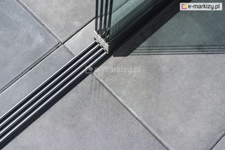 Zamocowana do kwadratowych płyt betonowych prowadnica dolna systemu Auris z czterema torami oraz czterema panelami szklanymi przesuwnymi.