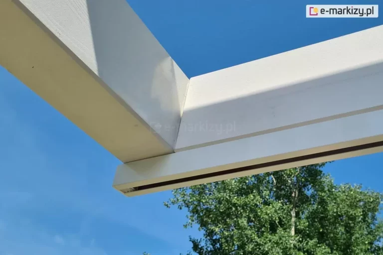 Narożnik pergoli drewnianej Moza pomalowanej na biało z zamocowanym do bocznej belki białym profilem prowadnicy dachu rzymskiego Supro. W tle widoczna wysoka brzoza z zielonymi liśćmi i błękitne niebo.