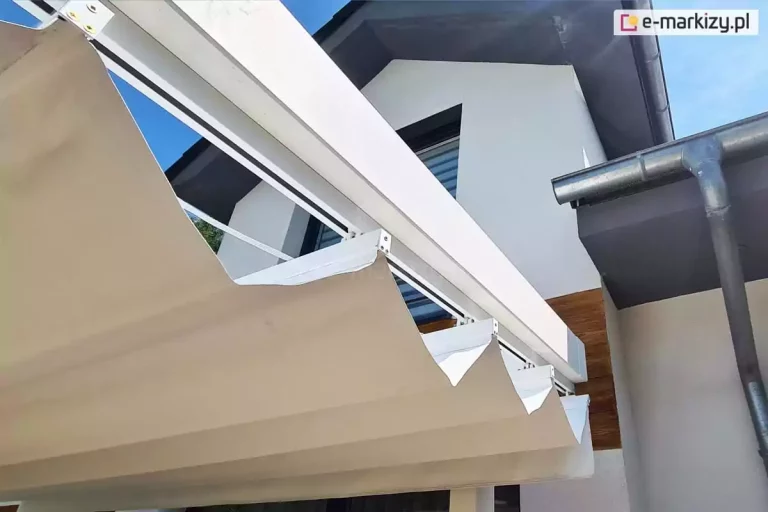 Widok na prowadnicę dachu Supro przykręconą do spodu belki konstrukcji pergoli drewnianej malowanej na kolor biały. W tle widoczne okno w wykuszu oraz rynna spadzistego dachu po prawej.