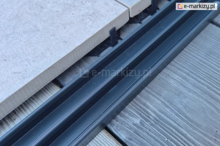 Zbliżenie antracytowej aluminiowej prowadnicy dolnej systemu szyb przesuwnych Miros z widocznymi trzema torami. Prowadnica zamocowana na powierzchni z szerokich desek z widocznym obok pokryciem tarasu grubymi płytami gresowymi w jasnym kolorze.