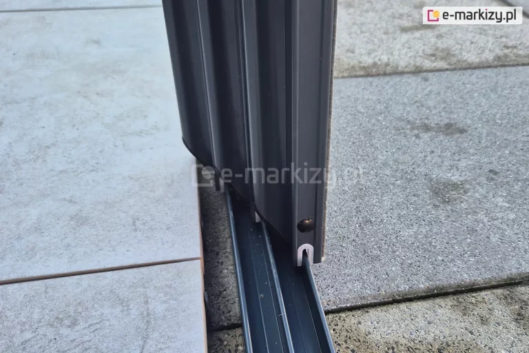 Zamocowana do płyt betonowych dolna prowadnica systemu szklanych drzwi przesuwnych ramowych Sotro z antracytowym wykończeniem i widoczną klamką. Po lewej przestrzeń tarasu wyłożona dodatkowymi płytkami dekoracyjnymi.