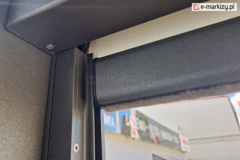 Słupek konstrukcji pergoli sb 400 Selt w kolorze antracytowym z zamontowaną refleksolą ziiip 120 umieszczoną w prowadnicy bocznej. Konstrukcja refleksola dopasowana została do koloru pergoli, w belce dolnej widoczna uszczelka szczotkowa oraz zwinięta tkanina screen w kolorze beżowym.