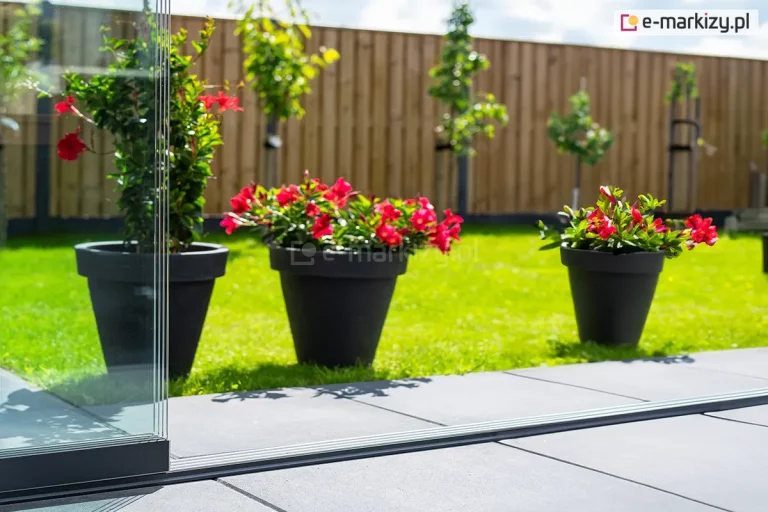 Detal dolnej prowadnicy systemu szklanych ścian przesuwnych Auris - Fiano Deponti na tle ogrodu. Na pierwszym planie widoczna jest krawędź czterech tafli szkła hartowanego, które są osadzone w czarnym profilu jezdnym. Szkło przesuwa się po niskiej, srebrnej szynie zamontowanej na płaskich, szarych płytach tarasowych. Drugi plan zajmuje zadbany trawnik, na którym ustawiono trzy czarne donice z kwitnącymi czerwonymi kwiatami. Całość ogrodu jest ogrodzona pionowym, drewnianym płotem z jasnych desek, a w oddali widoczne są młode drzewa i słoneczne, lekko zachmurzone niebo.