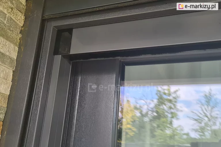 Zbliżenie narożnika moskitiery plisowanej Click Rol z widocznym czarnym łącznikiem profilu górnego i bocznego. Całość zamocowana do ramy okna tarasowego o antracytowym kolorze, pomiędzy ramą okna a prowadnicami rolety zewnętrznej.