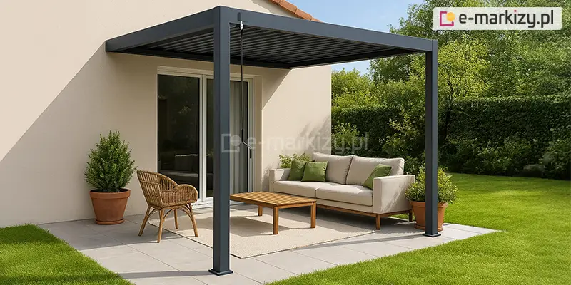Nowoczesna pergola tarasowa Marlo w kolorze antracytowym, zamontowana przyściennie do elewacji budynku o jasnym, beżowym wykończeniu. Konstrukcja zadaszenia opiera się na dwóch solidnych profilach przednich, a dach składa się z poziomych lameli ustawionych w pozycji zamkniętej. Pod pergolą zaaranżowano strefę wypoczynkową na tarasie wyłożonym jasnymi płytami, na którą składają się: jasnobeżowa sofa z zielonymi poduszkami, drewniany stolik kawowy, pleciony fotel oraz dywan zewnętrzny. Po bokach ustawiono rośliny w glinianych donicach, a całość otacza zadbany trawnik oraz gęsty, zielony żywopłot. Przy przednim słupie widoczna jest ręczna korba służąca do regulacji kąta nachylenia lameli.