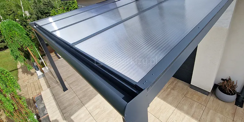 Widok z góry na rynnę przednią o zaokrąglonym profilu w zadaszeniu Lunaro o antracytowej konstrukcji. Widoczne łączenie krokwi dwachowych, paneli płyt poliwęglanu komorowego. Pod zadaszeniem widoczny taras wyłożony płytkami gresowymi o drewnopodobnym wzorze oraz zielone rośliny posadzone w ogrodzie.