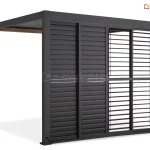 Antracytowa pergola Gloria z systemem przesuwnych paneli żaluzjowych, przedstawiona w ujęciu od strony frontu. Zabudowa składa się z czterech skrzydeł, które są przesunięte w prawą stronę, pozostawiając lewą część boku pergoli otwartą. Dwa panele po lewej stronie mają lamele ustawione w pozycji całkowicie zamkniętej, tworząc jednolitą osłonę. Dwa panele po prawej stronie mają lamele ustawione poziomo, co pozwala na swobodny przepływ światła i wgląd do wnętrza. Każde skrzydło posiada aluminiową ramę z poprzeczką dzielącą panel na dwie równe sekcje pionowe. Konstrukcja pergoli opiera się na ciemnych słupach przednich oraz tylnych słupach o strukturze imitującej drewno. W dolnej części lewego słupa widoczne jest gniazdo z podłączonym przewodem zasilającym.