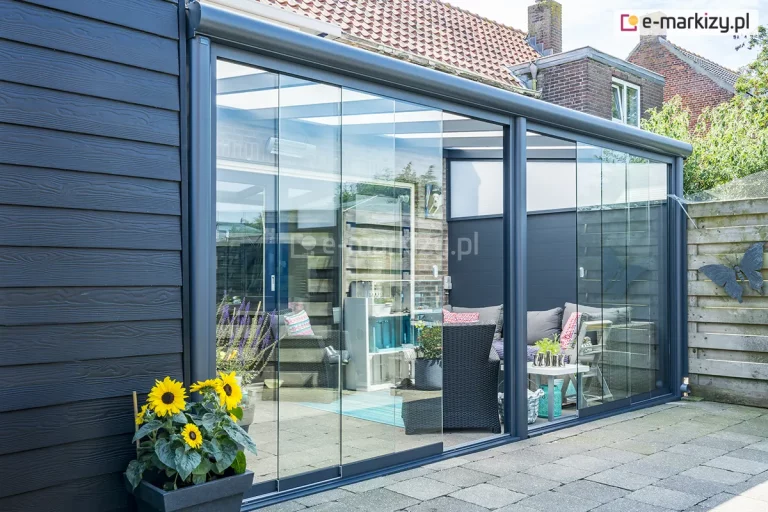 Zewnętrzne ujęcie nowoczesnego, w pełni zabudowanego zadaszenia tarasowego Lunaro, zamontowanego przy budynku o ciemnej, drewnianej elewacji. Frontową ścianę konstrukcji stanowią bezramowe, szklane panele przesuwne, które pozwalają na całkowite zamknięcie przestrzeni i stworzenie ogrodu zimowego. Wewnątrz widoczna jest przytulna strefa wypoczynkowa z szarymi meblami technorattanowymi, ozdobnymi poduszkami oraz błękitnym dywanem. Na pierwszym planie, na wybrukowanym podłożu, ustawiono donicę z jaskrawożółtymi słonecznikami, a w tle widać dachy sąsiednich domów z czerwonej dachówki.