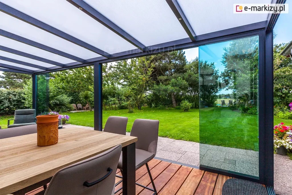 Widok spod zadaszenia tarasowego Lunaro (Bosco Deponti), wyposażonego w system szklanych ścian przesuwnych. Dach konstrukcji w kolorze antracytowym wypełniają białe panele z poliwęglanu komorowego, podtrzymywane przez aluminiowe krokwie z punktowym oświetleniem LED. Jedna z transparentnych szyb bocznych jest częściowo przesunięta, otwierając przejście na zielony, zadbany ogród z gęstą roślinnością i drzewami. Na tarasie, w strefie wyłożonej drewnianymi deskami, ustawiono nowoczesny stół z jasnym blatem oraz dopasowane szare krzesła.