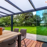 Widok spod zadaszenia tarasowego Lunaro (Bosco Deponti), wyposażonego w system szklanych ścian przesuwnych. Dach konstrukcji w kolorze antracytowym wypełniają białe panele z poliwęglanu komorowego, podtrzymywane przez aluminiowe krokwie z punktowym oświetleniem LED. Jedna z transparentnych szyb bocznych jest częściowo przesunięta, otwierając przejście na zielony, zadbany ogród z gęstą roślinnością i drzewami. Na tarasie, w strefie wyłożonej drewnianymi deskami, ustawiono nowoczesny stół z jasnym blatem oraz dopasowane szare krzesła.