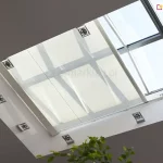 Widok od wewnątrz na duży, prostokątny świetlik dachowy z zainstalowaną pod nim wewnętrzną roletą poziomą (system typu Veranda) w kolorze białym lub kremowym. Półtransparentna tkanina rolety jest napięta na aluminiowych profilach i częściowo zasłania przeszklenie, rozpraszając wpadające światło słoneczne. Rama świetlika jest biała, zintegrowana z sufitem, na którym zamontowano punktowe, kwadratowe oprawy oświetleniowe LED rozmieszczone symetrycznie wokół wnęki okiennej. W prawym górnym rogu widoczny jest mechanizm siłownika do otwierania okna dachowego, a w dolnej części zdjęcia fragmenty zielonych liści rośliny doniczkowej.