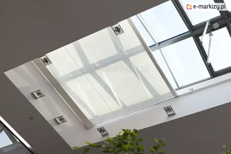 Widok od wewnątrz na duży, prostokątny świetlik dachowy z zainstalowaną pod nim wewnętrzną roletą poziomą (system typu Veranda) w kolorze białym lub kremowym. Półtransparentna tkanina rolety jest napięta na aluminiowych profilach i częściowo zasłania przeszklenie, rozpraszając wpadające światło słoneczne. Rama świetlika jest biała, zintegrowana z sufitem, na którym zamontowano punktowe, kwadratowe oprawy oświetleniowe LED rozmieszczone symetrycznie wokół wnęki okiennej. W prawym górnym rogu widoczny jest mechanizm siłownika do otwierania okna dachowego, a w dolnej części zdjęcia fragmenty zielonych liści rośliny doniczkowej.