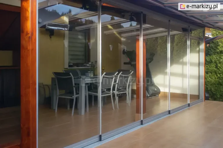 Widok na nowoczesną, przeszkloną zabudowę tarasu z przezroczystymi, przesuwanymi panelami szklanymi osadzonymi w cienkich, aluminiowych prowadnicach. Wewnątrz zadaszonej przestrzeni znajduje się prostokątny stół ogrodowy z sześcioma krzesłami oraz urządzenie do ćwiczeń – orbitrek. Na ścianie zamontowany jest promiennik ciepła. Konstrukcja oparta na drewnianych słupach, z dachem z przezroczystego materiału. Po prawej stronie widoczne są wysokie, gęste tuje stanowiące zieloną osłonę. Taras wyłożony jest płytkami w odcieniu ciepłego brązu.