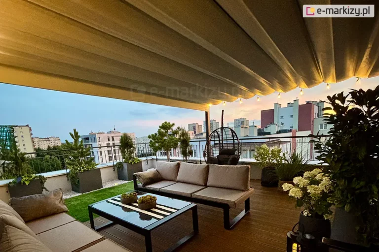 Przestronna strefa wypoczynkowa zaaranżowana na tarasie dachowym, osłoniętą dużą, poziomą roletą rzymską Supro z tkaniną Opatex w kolorze beżowym. Tkanina zadaszenia jest pofalowana i zamontowana na prowadnicach, a wzdłuż jej krawędzi rozwieszono girlandę świetlną z małymi punktami świetlnymi o ciepłej barwie. Wyposażenie tarasu obejmuje nowoczesne meble ogrodowe: szarą sofę o prostej, ciemnej konstrukcji z miękkimi poduchami oraz niski stolik kawowy z ciemnym blatem, na którym ustawiono dekoracyjne misy z roślinnością. Nawierzchnia tarasu jest dwudzielna – część pokrywają brązowe deski kompozytowe, a fragment przy balustradzie wyłożono sztuczną trawą oraz jasnym żwirem dekoracyjnym. Wzdłuż metalowej, czarnej barierki ustawiono liczne rośliny w donicach, w tym formowane iglaki, krzewy oraz kwitnące na biało hortensje. W tle widoczna jest panorama miasta z wielorodzinną zabudową mieszkalną pod jasnym, wieczornym niebem z delikatnymi akcentami zachodzącego słońca.
