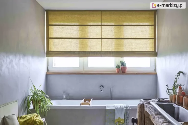 Zdjęcie przedstawia minimalistyczną i surową łazienkę z oknem przesłoniętym rzymską roletą w kolorze musztardowym lub oliwkowej zieleni. Tkanina rolety ma wyraźną strukturę grubego splotu przypominającego naturalny len i jest półtransparentna, dzięki czemu przepuszcza do wnętrza miękkie, rozproszone światło. Roleta posiada regularne, poziome przeszycia z fiszbinami oraz pionowe wzmocnienie przebiegające przez środek, co dzieli jej powierzchnię na symetryczne prostokąty. Osłona jest podciągnięta do połowy wysokości okna, odsłaniając białą ramę trójskrzydłowego okna i drewniany parapet, na którym ustawiono dwie małe doniczki z kaktusami. Wnętrze łazienki wykończone jest betonem lub tynkiem strukturalnym w chłodnym, szarym odcieniu. Pod oknem znajduje się wbudowana, biała wanna o prostokątnym kształcie. Na jej krawędzi leży drewniana półka wannowa oraz przewieszony jest jasny ręcznik z żółtym, dekoracyjnym chwostem. Po lewej stronie wanny, na podłodze, stoi donica z zieloną rośliną o długich, wąskich liściach (zielistka), a obok niej widać fragment krzesła z przewieszoną żółtą tkaniną. Po prawej stronie kadru znajduje się rustykalna szafka podumywalkowa wykonana z grubego, surowego drewna z zamontowaną metalową umywalką. Na blacie obok umywalki ustawione są gliniane kubki i drobne akcesoria łazienkowe. Całość aranżacji łączy surowość szarych ścian z ciepłymi akcentami drewna i tkaniny, tworząc przestrzeń w stylu nowoczesnego wabi-sabi.