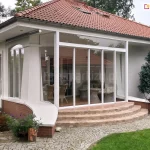 Nowoczesna zabudowa tarasu wykonana w formie przeszklonego ogrodu zimowego przy parterowym domu jednorodzinnym. Głównym elementem konstrukcyjnym są przesuwne drzwi szklane oparte na białych profilach aluminiowych, które tworzą transparentną ścianę frontową oraz boczną. System składa się z kilku pionowych paneli szklanych umieszczonych na szynach, co pozwala na szerokie otwarcie przestrzeni na ogród. Zabudowa została wkomponowana w bryłę budynku pod istniejącym zadaszeniem dachu pokrytego czerwoną dachówką ceramiczną. Konstrukcja opiera się na niskim podmurówce wykończonej czerwoną cegłą klinkierową, do której prowadzą półkoliste, szerokie schody wykonane z jasnej kostki brukowej. Wewnątrz przeszklonego pomieszczenia widoczne są meble ogrodowe z technorattanu, w tym fotele i stolik, oraz drewniana podłoga. Ściany domu są otynkowane na biało, a wokół tarasu znajduje się zadbany trawnik, krzewy ozdobne oraz ścieżka z szarej kostki granitowej. Po lewej stronie widoczna jest jednostka zewnętrzna klimatyzacji zamontowana na elewacji.