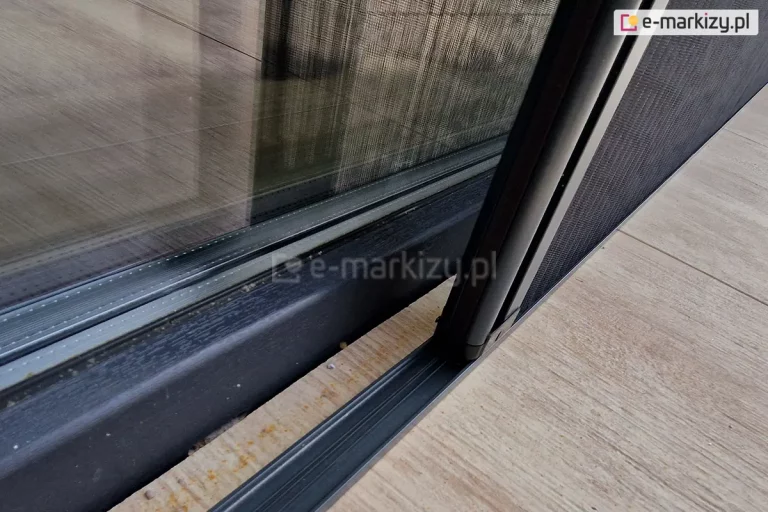 Zbliżenie niskiego 8mm progu dolnego moskitiery plisowanej Top Zag z widocznymi wewnątrz sznurkami, profilem bocznym siatki i częściowo rozsuniętą szara siatką. Próg naklejony na płytki gresowe o drewnianym wzorze w pobliżu antracytowej ramy okna tarasowego.