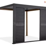 Antracytowa pergola Gloria z systemem przesuwnych shuttersów, zamontowanych na jednym z boków konstrukcji. Na zdjęciu widoczne są dwa panele żaluzjowe rozsuniecie na boki, które tworzą szerokie światło przejścia w środkowej części. Każdy panel składa się z dwóch sekcji wypełnionych poziomymi, nieruchomymi lamelami o szerokim profilu, które zapewniają ochronę przed słońcem i prywatność. Rama pergoli oraz panele przesuwne są wykonane z aluminium w kolorze ciemnoantracytowym, natomiast dwa tylne słupy konstrukcyjne mają wykończenie w kolorze naturalnego drewna. W lewym dolnym rogu przedniego słupa widoczne jest wyprowadzenie przewodu elektrycznego. Całość osadzona jest na stabilnych stopach montażowych, a dach konstrukcji tworzą dopasowane kolorystycznie lamele.