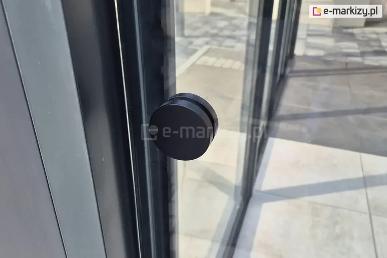 Zbliżenie na okrągły, czarny uchwyt zamontowany na tafli przesuwnego panelu szklanego w bezramowej zabudowie tarasu Miros. Uchwyt ma prostą, matową powierzchnię i zamocowany jest centralnie na wysokości ręki. W tle widoczna posadzka z dużych płyt gresowych w odcieniach szarości oraz szklane ściany konstrukcji z aluminiowymi profilami w kolorze antracytowym. Zdjęcie wykonano w świetle dziennym, na zewnątrz nowoczesnego budynku.