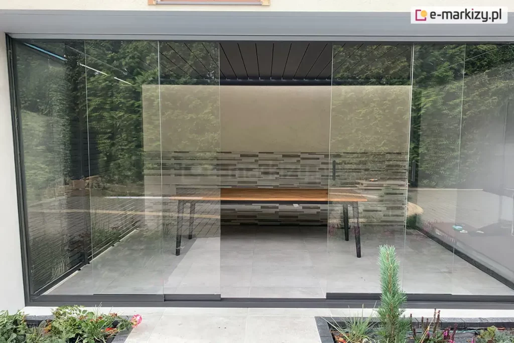 Zdjęcie przedstawia nowoczesną pergolę aluminiową Rubio w ciemnoszarym kolorze, która została zabudowana przesuwnym systemem szklanych paneli. Bezramowe tafle szkła są zamontowane na dolnej i górnej prowadnicy, tworząc przezroczystą ścianę frontową, przez którą widoczne jest wnętrze tarasu z drewnianym stołem na czarnych nogach. W tle znajduje się dekoracyjna ściana o strukturze kamiennych płytek w odcieniach szarości i beżu, a powyżej widoczne jest zadaszenie z ciemnych lameli. Cała konstrukcja jest osadzona na jasnoszarym tarasie z płyt wielkoformatowych, otoczonym zadbaną roślinnością ogrodową, w tym iglakami i niskimi krzewami.