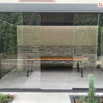 Zdjęcie przedstawia nowoczesną pergolę aluminiową Rubio w ciemnoszarym kolorze, która została zabudowana przesuwnym systemem szklanych paneli. Bezramowe tafle szkła są zamontowane na dolnej i górnej prowadnicy, tworząc przezroczystą ścianę frontową, przez którą widoczne jest wnętrze tarasu z drewnianym stołem na czarnych nogach. W tle znajduje się dekoracyjna ściana o strukturze kamiennych płytek w odcieniach szarości i beżu, a powyżej widoczne jest zadaszenie z ciemnych lameli. Cała konstrukcja jest osadzona na jasnoszarym tarasie z płyt wielkoformatowych, otoczonym zadbaną roślinnością ogrodową, w tym iglakami i niskimi krzewami.