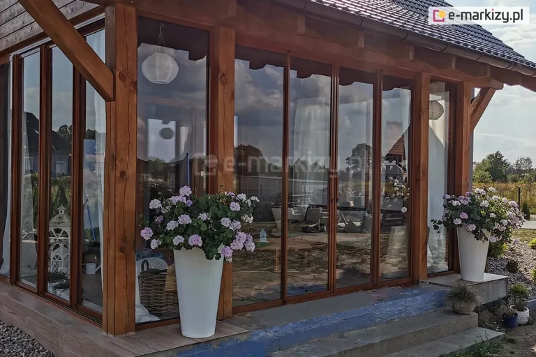 System szklanych ścian przesuwnych Sotro zainstalowany w klasycznej, drewnianej konstrukcji zadaszenia ogrodowego. Panele szklane zostały osadzone w aluminiowych profilach w kolorze brązowym, który harmonizuje z naturalnym odcieniem belek nośnych i słupów. Zabudowa zamyka przestrzeń tarasu, tworząc szczelną barierę przed warunkami atmosferycznymi, co potwierdzają wyraźne odbicia nieba i chmur na powierzchni szyb. Przed szklanymi ścianami ustawione są duże, białe donice z kwitnącymi roślinami, a wewnątrz widoczne są elementy dekoracyjne, takie jak papierowe lampiony i białe lampiony ogrodowe. Całość osadzona jest na podwyższonej betonowej podstawie wykończonej płytami, co zapewnia stabilność systemu przesuwnego w tradycyjnej architekturze ogrodowej.