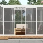 Zdjęcie prezentuje białą, aluminiową konstrukcję pergoli ogrodowej Gloria z systemem przesuwnych ścian bocznych, wypełnionych przeziernym poliwęglanem. Frontowa ściana składa się z czterech paneli ujętych w białe ramy z poprzecznymi szprosami, przy czym dwa środkowe moduły są rozsunięte na boki, tworząc otwarte przejście do wnętrza. Zastosowany poliwęglan ma wyraźne, ciemnoszare zabarwienie, które zapewnia prywatność i ochronę przed słońcem, jednocześnie pozwalając na wgląd do środka, gdzie widoczna jest sofa z jasnymi poduszkami oraz drewniany ławostół. Konstrukcja zadaszenia jest płaska, wyposażona w system ruchomych lameli dachowych w kolorze białym. Pergola została ustawiona na tarasie z jasnobrązowych desek, graniczącym z pasem dekoracyjnego szarego żwiru i dużymi płytami chodnikowymi. W tle widoczny jest gęsty las iglasty pod czystym, błękitnym niebem, co podkreśla nowoczesny i funkcjonalny charakter zabudowy wkomponowanej w naturalne otoczenie.