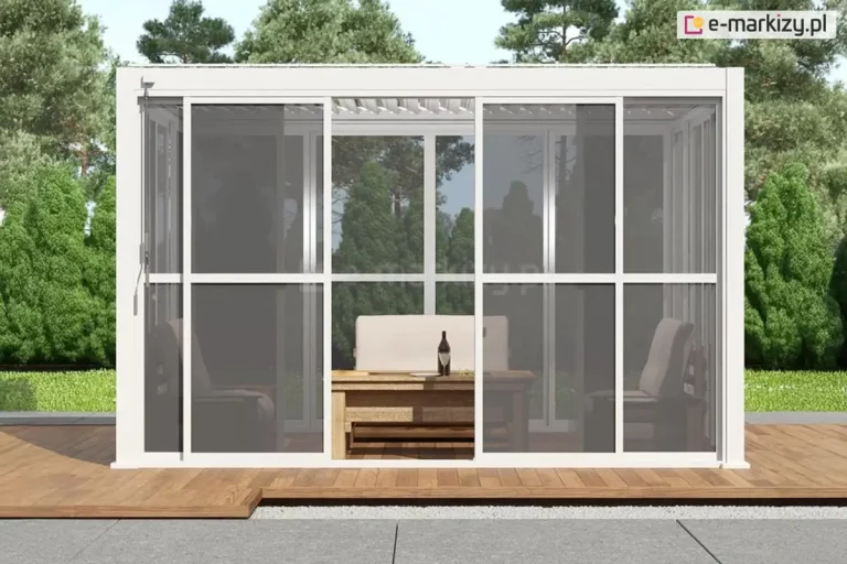 Zdjęcie prezentuje białą, aluminiową konstrukcję pergoli ogrodowej Gloria z systemem przesuwnych ścian bocznych, wypełnionych przeziernym poliwęglanem. Frontowa ściana składa się z czterech paneli ujętych w białe ramy z poprzecznymi szprosami, przy czym dwa środkowe moduły są rozsunięte na boki, tworząc otwarte przejście do wnętrza. Zastosowany poliwęglan ma wyraźne, ciemnoszare zabarwienie, które zapewnia prywatność i ochronę przed słońcem, jednocześnie pozwalając na wgląd do środka, gdzie widoczna jest sofa z jasnymi poduszkami oraz drewniany ławostół. Konstrukcja zadaszenia jest płaska, wyposażona w system ruchomych lameli dachowych w kolorze białym. Pergola została ustawiona na tarasie z jasnobrązowych desek, graniczącym z pasem dekoracyjnego szarego żwiru i dużymi płytami chodnikowymi. W tle widoczny jest gęsty las iglasty pod czystym, błękitnym niebem, co podkreśla nowoczesny i funkcjonalny charakter zabudowy wkomponowanej w naturalne otoczenie.