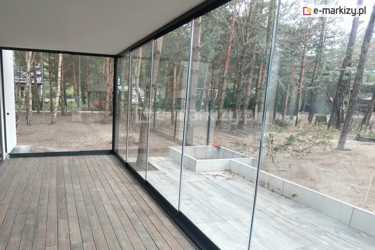 Przeszklona weranda lub zabudowa tarasowa z systemem bezramowych szyb przesuwnych Miros, zamontowana przy nowoczesnym budynku zlokalizowanym w lesie sosnowo-brzozowym. Wnętrze wyłożone drewnianą podłogą z ciemnych desek, otoczone przezroczystymi taflami szkła osadzonymi w czarnych prowadnicach dolnych i górnych. Na zewnątrz widoczne są drzewa, ogrodzenie z siatki oraz betonowe donice lub rabaty ogrodowe w zabudowie narożnej. Naturalne światło przenika przez szyby, tworząc spokojną, leśną atmosferę.