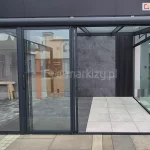 frontowe ujęcie systemowej zabudowy szklanej Sotro zintegrowanej z konstrukcją pergoli o ciemnym wykończeniu. Centralnym elementem jest częściowo rozsunięty system paneli szklanych, który ujawnia wielotorową prowadnicę dolną oraz mechanizm nakładania się na siebie poszczególnych skrzydeł. Wnętrze konstrukcji wykończone jest dekoracyjną ścianą z ciemnoszarych płyt o teksturze kamienia oraz pionowymi, czarnymi lamelami, co podkreśla nowoczesny design instalacji. Podłoga wewnątrz podzielona jest na strefę z ciemnych i jasnych płyt wielkoformatowych, tworząc geometryczny kontrast. Przez szklane ściany boczne i frontowe widoczne jest otoczenie miejskie oraz inne elementy architektury ogrodowej, co obrazuje wysoką transparentność zastosowanego szkła bezpiecznego i lekkość optyczną profili aluminiowych.