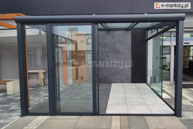 frontowe ujęcie systemowej zabudowy szklanej Sotro zintegrowanej z konstrukcją pergoli o ciemnym wykończeniu. Centralnym elementem jest częściowo rozsunięty system paneli szklanych, który ujawnia wielotorową prowadnicę dolną oraz mechanizm nakładania się na siebie poszczególnych skrzydeł. Wnętrze konstrukcji wykończone jest dekoracyjną ścianą z ciemnoszarych płyt o teksturze kamienia oraz pionowymi, czarnymi lamelami, co podkreśla nowoczesny design instalacji. Podłoga wewnątrz podzielona jest na strefę z ciemnych i jasnych płyt wielkoformatowych, tworząc geometryczny kontrast. Przez szklane ściany boczne i frontowe widoczne jest otoczenie miejskie oraz inne elementy architektury ogrodowej, co obrazuje wysoką transparentność zastosowanego szkła bezpiecznego i lekkość optyczną profili aluminiowych.