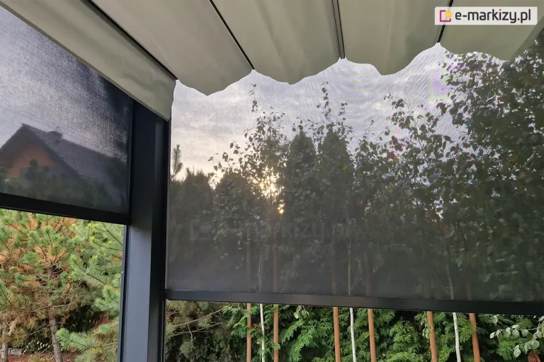 Pergola Tako z antracytowa konstrukcja i białym dachem rzymskim oraz osłoniętymi roletami screen bocznymi ścianami. Opuszczone częściowo osłony boczne do pergoli przepuszczają światło chroniąc przed intensywnym słońcem widocznym pomiędzy gałęziami drzew otaczających pergolę.