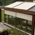 Przeszklone patio nowoczesnego domu z roletami zewnętrznymi typu ZIP L zamontowanymi na dużych, drewnianych ramach okiennych. Rolety w jasnym, beżowym kolorze osłaniają wnętrze ogrodu zimowego, zapewniając prywatność i ochronę przed słońcem. Konstrukcja dachu wykonana z drewna łączy się z fragmentem dachu pokrytego grafitową dachówką. Przeszkloną część dachu osłonięto roletami poziomymi w kolorystyce dopasowanej do stylu i barw domu. Wewnątrz pomieszczenia widoczne są rośliny doniczkowe oraz zestaw wypoczynkowy z kanapą i stolikiem. Otoczenie domu stanowi zadbany trawnik, roślinność ozdobna i alejka z kamienną nawierzchnią prowadząca przez ogród.