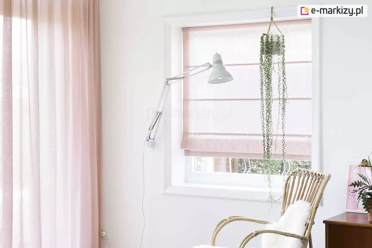 Zdjęcie przedstawia jasne, kobiece, dziewczęce, wnętrze utrzymane w pastelowej, biało-różowej kolorystyce, w którym głównym elementem jest dekoracja okienna. W centralnej części znajduje się kwadratowe okno z białą ramą, przesłonięte rzymską roletą wykonaną z transparentnego, jasnoróżowego materiału o delikatnym splotu. Roleta jest częściowo podciągnięta, a jej powierzchnia jest podzielona poziomymi fiszbinami na równe sekcje, przy czym dolna część tworzy estetyczną zakładkę. Przez półprzezroczystą tkaninę wpada do środka miękkie, dzienne światło. Po lewej stronie okna, na ścianie, zamontowana jest długa, biała lampa kreślarska na ruchomym wysięgniku, której klosz skierowany jest w stronę centrum pokoju. Przy górnej krawędzi okna wisi biała doniczka z rośliną o bardzo długich, cienkich pędach z drobnymi listkami (starzec zwisający), które opadają pionowo w dół, sięgając niemal do połowy wysokości wnęki okiennej. Po lewej stronie kadru widoczny jest fragment długiej, zwiewnej zasłony w tym samym odcieniu pudrowego różu co roleta, co tworzy spójną aranżację tekstylną. W prawym dolnym rogu zdjęcia znajduje się fragment plecionego, rattanowego fotela o ażurowej strukturze, na którym leży białe, puszyste futro. Obok fotela widać róg ciemnobrązowej, drewnianej komody, na której stoi mała roślina doniczkowa oraz różowa grafika w ramce. Ściany pomieszczenia są białe i gładkie, co podkreśla lekkość i romantyczny charakter wnętrza. Całość kompozycji jest niezwykle świetlista i przytulna.