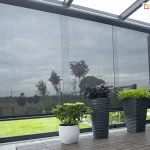 Nowoczesna pergola ogrodowa z przeszklonymi ścianami i zamontowanymi roletami Refleksol 103 Selt w eleganckim, grafitowym kolorze. Półprzezroczyste rolety tworzą subtelną barierę między wnętrzem a ogrodem, chroniąc przed słońcem i zapewniając intymność. Na drewnianej podłodze ustawiono cztery ozdobne donice z roślinami — białą ceramiczną oraz trzy antracytowe, o falistej strukturze, z różnorodną zielenią i bordowymi liśćmi. Rośliny harmonizują kolorystycznie z nowoczesnym stylem konstrukcji. Całość tworzy nowoczesną, funkcjonalną i stylową przestrzeń relaksu przy domu.