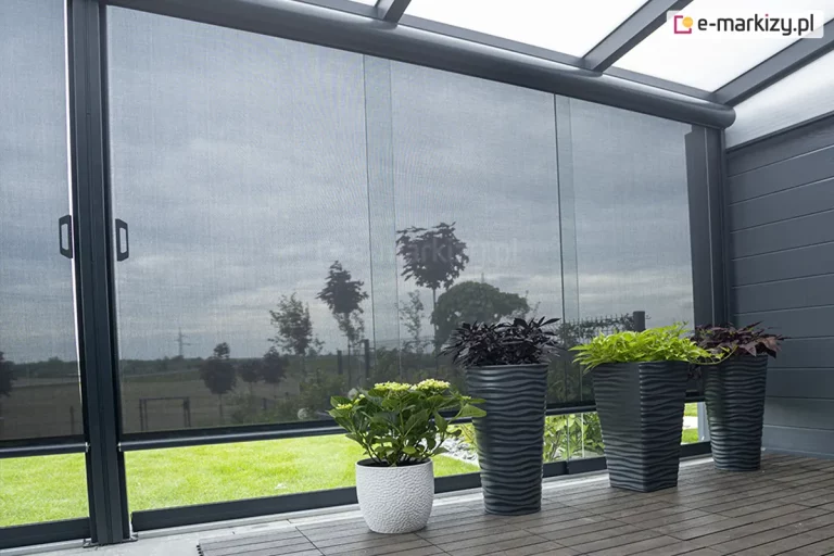 Nowoczesna pergola ogrodowa z przeszklonymi ścianami i zamontowanymi roletami Refleksol 103 Selt w eleganckim, grafitowym kolorze. Półprzezroczyste rolety tworzą subtelną barierę między wnętrzem a ogrodem, chroniąc przed słońcem i zapewniając intymność. Na drewnianej podłodze ustawiono cztery ozdobne donice z roślinami — białą ceramiczną oraz trzy antracytowe, o falistej strukturze, z różnorodną zielenią i bordowymi liśćmi. Rośliny harmonizują kolorystycznie z nowoczesnym stylem konstrukcji. Całość tworzy nowoczesną, funkcjonalną i stylową przestrzeń relaksu przy domu.