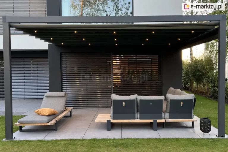Nowoczesny, zadaszony taras przy eleganckim domu jednorodzinnym. Ujęcie frontalne ukazuje symetryczną kompozycję przestrzeni. Taras przykryty jest ciemnoszarą pergolą o prostej, aluminiowej konstrukcji z belkowaniem w dachu, w którym zamontowano kilkanaście drobnych punktów świetlnych LED o ciepłej barwie – aktualnie włączonych. Pod zadaszeniem ustawiono dwa elementy zestawu wypoczynkowego: po lewej stronie – pojedynczy, nowoczesny leżak z jasnoszarą tapicerką i regulowanym zagłówkiem, na którym spoczywa poduszka dekoracyjna w kolorze ciemnej żółci; po prawej – duża, narożna sofa ogrodowa w tej samej stylistyce i kolorystyce, złożona z kilku segmentów ustawionych w literę „L”. Meble posiadają drewniane podstawy i czarne, metalowe nogi. W tle widoczna jest elewacja budynku w dwóch kontrastujących odcieniach – jasnym i grafitowym. Całą ścianę wypełniają szerokie przesuwne okna lub drzwi tarasowe zasłonięte poziomymi, zewnętrznymi żaluzjami. Po prawej stronie znajduje się niski, czarny stolik w formie ażurowego kosza. Taras wyłożony jest dużymi, prostokątnymi płytkami w kolorze jasnoszarym i otoczony starannie utrzymanym, zielonym trawnikiem. W tle, po bokach, widać ogrodową zieleń: liczne krzewy, drzewka i drzewa liściaste.