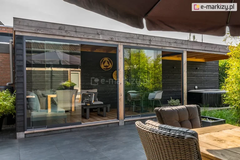 Nowoczesna przybudówka ogrodowa o konstrukcji hybrydowej, wyposażona w system bezramowych szklanych drzwi przesuwnych Auris. Zdjęcie przedstawia szeroki front wolnostojącego ogrodu letniego, gdzie panele szklane osadzone są w antracytowych profilach dolnych. Drzwi są częściowo rozsuniecie, otwierając widok na eleganckie, ciemne wnętrze z drewnianym sufitem i szarymi meblami wypoczynkowymi. Konstrukcja łączy naturalne drewniane słupy z czarnymi, poziomymi deskami elewacyjnymi. Na zewnątrz znajduje się taras z ciemnych płyt betonowych, a w tle widoczne jest jacuzzi oraz bujna zieleń ogrodowa (bambusy). Całość tworzy luksusową strefę relaksu, w której system Auris zapewnia ochronę przed wiatrem bez utraty panoramicznego widoku.