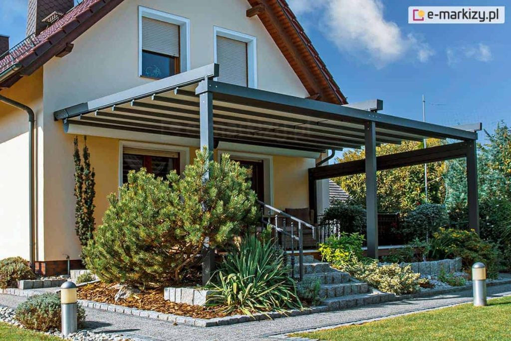 nowoczesna pergola aluminiowa Solid zamontowana na tarasie przy domu jednorodzinnym. Konstrukcja w kolorze ciemnego antracytu wspiera dach tkaninowy, który jest całkowicie rozwinięty, chroniąc przestrzeń tarasu przed słońcem. Budynek posiada jasną, beżową elewację, brązową dachówkę oraz okna z białymi ramami i zewnętrznymi roletami. Taras jest podniesiony względem ogrodu, a prowadzą do niego kamienne schody otoczone starannie zaplanowaną roślinnością, w tym dużym krzewem iglastym (sosną) oraz ozdobną jukką. Na pierwszym planie widoczny jest fragment trawnika oraz ogrodowe lampy słupkowe wzdłuż ścieżki wyłożonej grafitowymi płytami chodnikowymi.