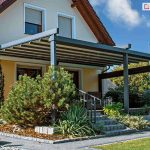 nowoczesna pergola aluminiowa Solid zamontowana na tarasie przy domu jednorodzinnym. Konstrukcja w kolorze ciemnego antracytu wspiera dach tkaninowy, który jest całkowicie rozwinięty, chroniąc przestrzeń tarasu przed słońcem. Budynek posiada jasną, beżową elewację, brązową dachówkę oraz okna z białymi ramami i zewnętrznymi roletami. Taras jest podniesiony względem ogrodu, a prowadzą do niego kamienne schody otoczone starannie zaplanowaną roślinnością, w tym dużym krzewem iglastym (sosną) oraz ozdobną jukką. Na pierwszym planie widoczny jest fragment trawnika oraz ogrodowe lampy słupkowe wzdłuż ścieżki wyłożonej grafitowymi płytami chodnikowymi.